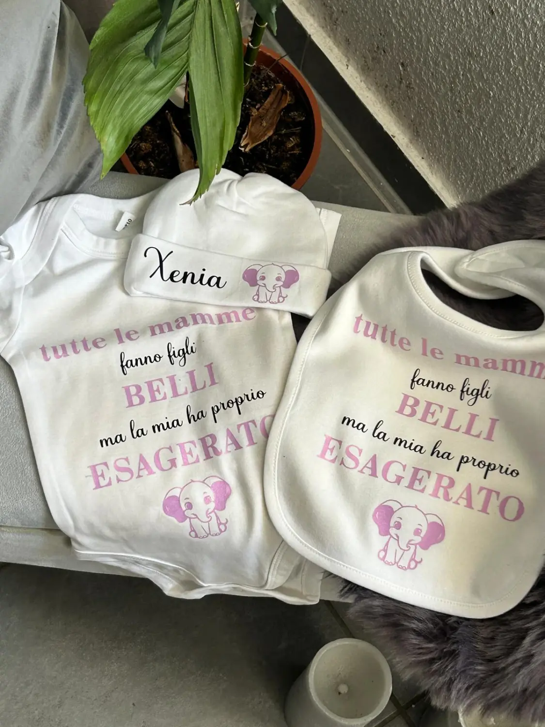 Ein personalisiertes Baby-Geschenkset mit Strampler, Mütze und Lätzchen. Alle Sets tragen den Namen „Xenia“ und ein verspieltes Elefantenmotiv. Dazu der italienische Spruch: „Tutte le mamme fanno figli BELLI ma la mia ha proprio ESAGERATO“ von Marika’s Dream World